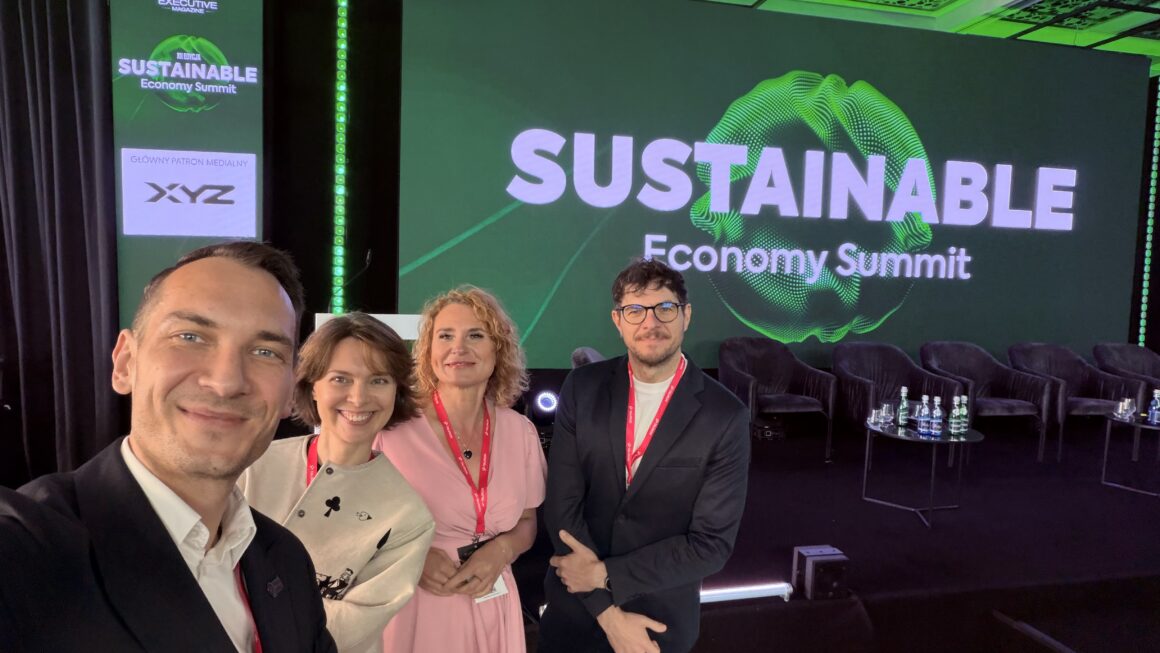 Cztery osoby stoją przed sceną konferencyjną podczas Sustainable Economy Summit i uśmiechają się do wspólnego zdjęcia. W tle widoczny jest duży ekran z nazwą wydarzenia oraz przygotowana scena z fotelami i stolikami dla panelistów. Zdjęcie pokazuje uczestników konferencji w przestrzeni wydarzenia poświęconego zrównoważonej gospodarce.