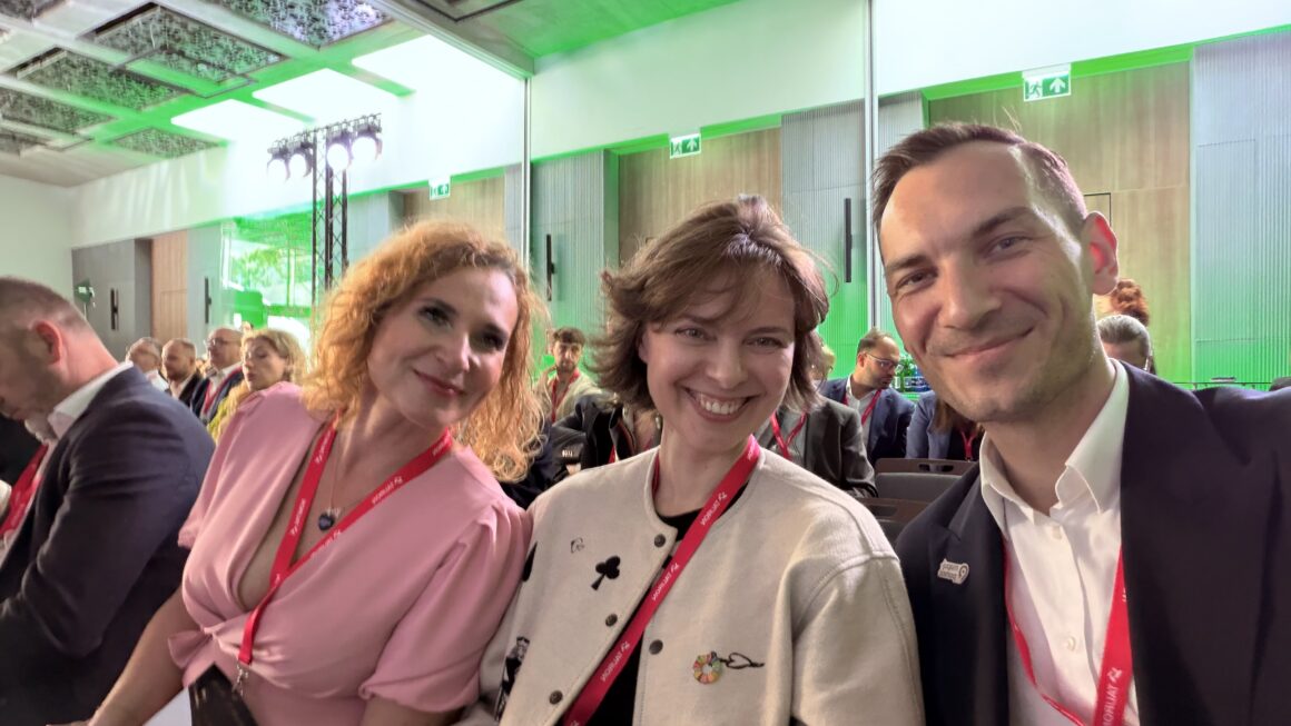 Trzy osoby siedzą na widowni podczas Sustainable Economy Summit i uśmiechają się do zdjęcia. W tle widoczna jest sala konferencyjna z uczestnikami wydarzenia oraz zielonym oświetleniem sceny. Zdjęcie pokazuje uczestników konferencji w trakcie wydarzenia poświęconego zrównoważonej gospodarce i ESG.
