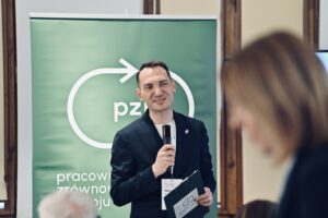 Prowadzący wydarzenie mówi do uczestników forum, stojąc przed zielonym banerem Pracowni Zrównoważonego Rozwoju i trzymając mikrofon oraz notatki.