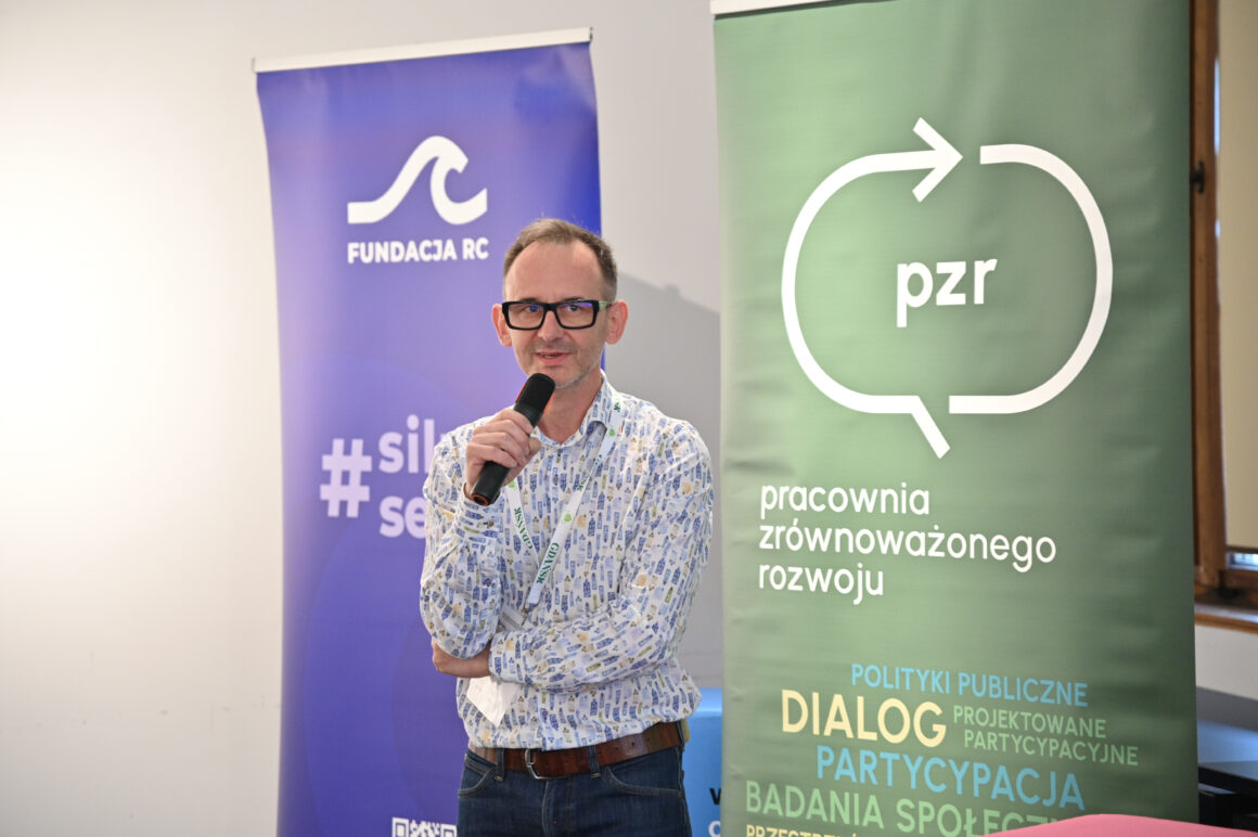 Mężczyzna w okularach stoi z mikrofonem w dłoni i przemawia do uczestników spotkania. Za nim znajdują się dwa roll-upy organizacji współtworzących wydarzenie: Fundacji RC oraz Pracowni Zrównoważonego Rozwoju. Na zielonym banerze PZR widoczne jest logo organizacji oraz hasła związane z dialogiem, partycypacją i badaniami społecznymi. Prelegent ma swobodną postawę i zwraca się bezpośrednio do publiczności, tworząc atmosferę otwartej rozmowy i współpracy. Zdjęcie przedstawia moment otwarcia lub prowadzenia części merytorycznej spotkania poświęconego partycypacji i dialogowi społecznemu.