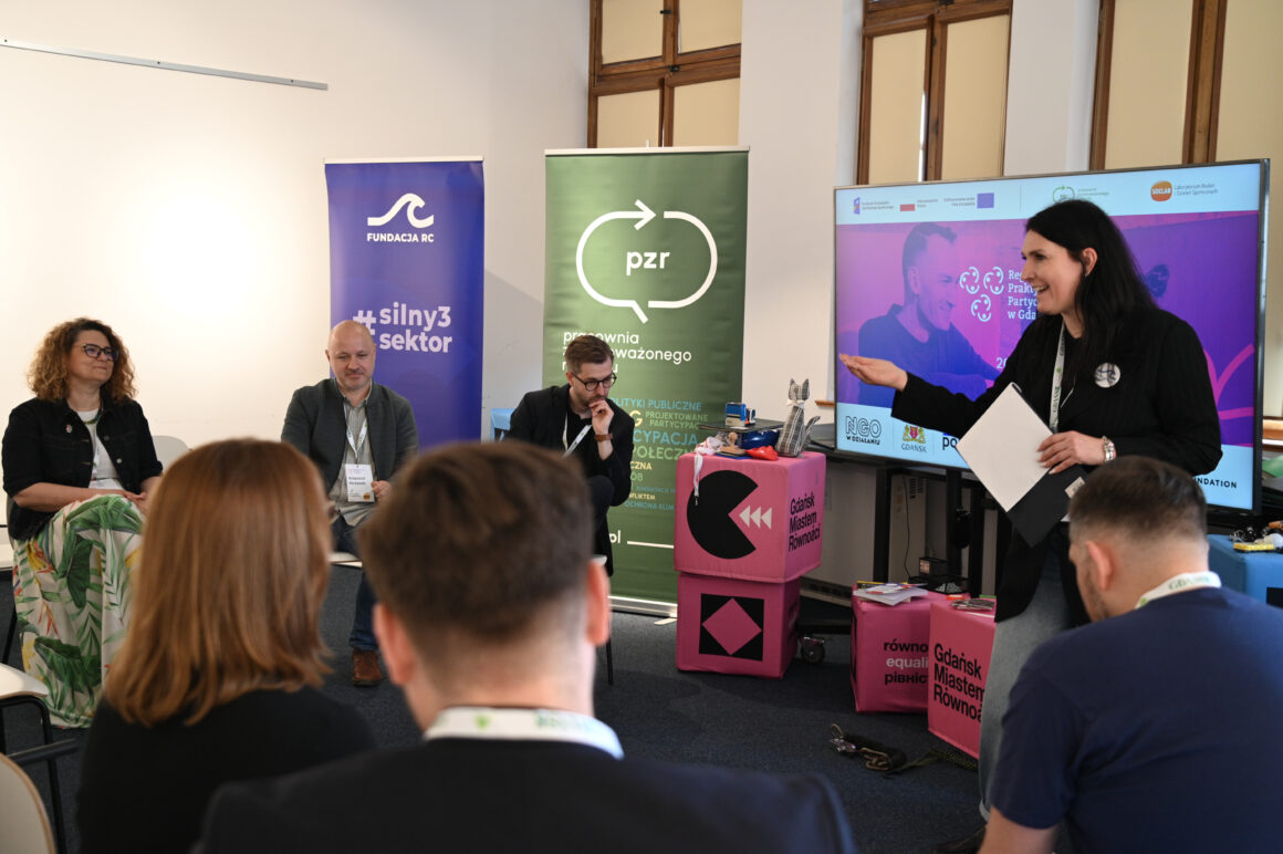 Magdalena Jabłońska prowadzi panel podczas Regionalnego Forum Praktyków Partycypacji w Gdańsku. Uczestnicy siedzą w półokręgu i słuchają dyskusji o liderstwie, partycypacji i zmianie kultury instytucji. W tle widoczne są bannery PZR i Fundacji RC oraz ekran z grafiką Forum.