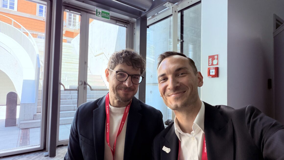 Selfie dwóch mężczyzn podczas konferencji Sustainable Economy Summit. Obaj uśmiechają się do aparatu i mają na szyjach czerwone identyfikatory konferencyjne. Siedzą przy przeszklonym wejściu do budynku, przez które widać schody i fragment nowoczesnej architektury na zewnątrz.