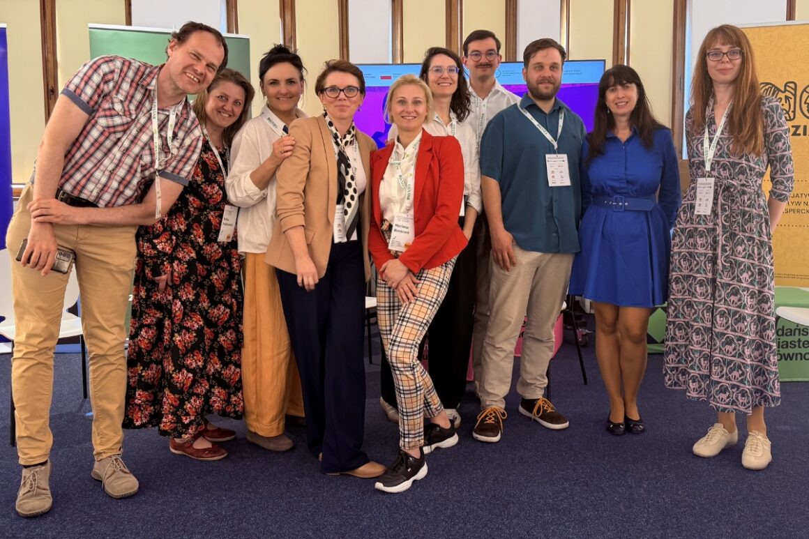 Zdjęcie grupowe uczestników Regionalnego Forum Praktyków Partycypacji. Dziewięć osób stoi razem i uśmiecha się do aparatu. W tle widoczne są ekrany oraz banery organizatorów wydarzenia. Fotografia pokazuje nieformalną atmosferę współpracy i integracji uczestników forum.