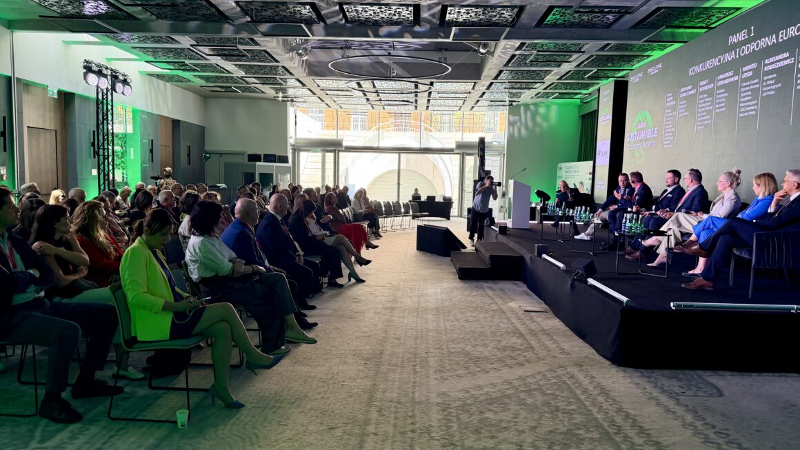 Szerokie ujęcie sali konferencyjnej podczas Sustainable Economy Summit. Na scenie trwa panel dyskusyjny dotyczący konkurencyjnej i odpornej Europy, a publiczność słucha rozmowy z miejsc dla uczestników. W tle widoczne są duże ekrany z nazwą wydarzenia i programem panelu oraz zielone oświetlenie sceny.
