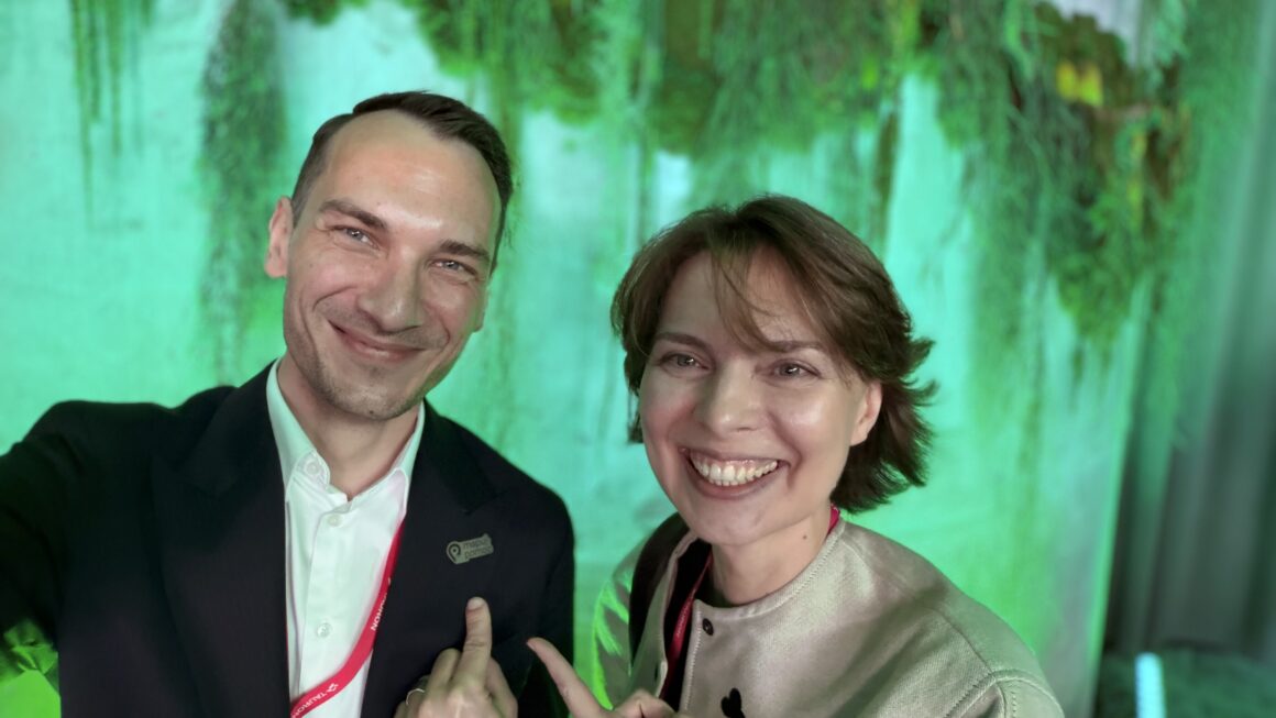 Selfie dwóch osób podczas konferencji Sustainable Economy Summit. Kobieta i mężczyzna uśmiechają się do aparatu na tle zielonej instalacji roślinnej i nastrojowego oświetlenia. Oboje mają na szyjach czerwone identyfikatory konferencyjne. Mężczyzna wskazuje dłonią na przypinkę przypiętą do marynarki.