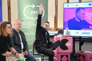 Uczestnik panelu unosi wysoko jedną rękę w geście zgłaszania się do odpowiedzi.
