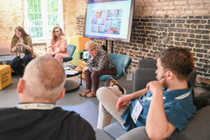 Panel dyskusyjny z udziałem osób siedzących na fotelach i kanapach i rozmawiających o miejscach wspólnych. W tle ceglana ściana oraz ekran ustawiony przy niej z wyświetlona prezentacją.