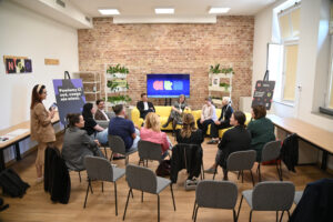 Sala warsztatowa podczas spotkania lub panelu dyskusyjnego. Uczestnicy siedzą w kręgu i rozmawiają w kameralnej atmosferze. W tle widać ekran z kolorowym logo „a4n”, ceglaną ścianę oraz stojące banery z hasłem „Powiemy Ci coś, czego nie wiesz”.
