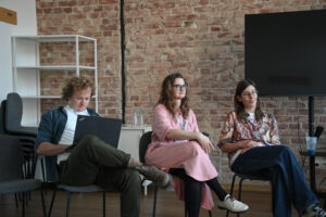 Trójka osób siedzących na krzesłach przysłuchuje się dyskusji podczas panelu o dostępności. W tle widać ceglaną ścianę. Mężczyzna z lewej strony pracuje na laptopie.