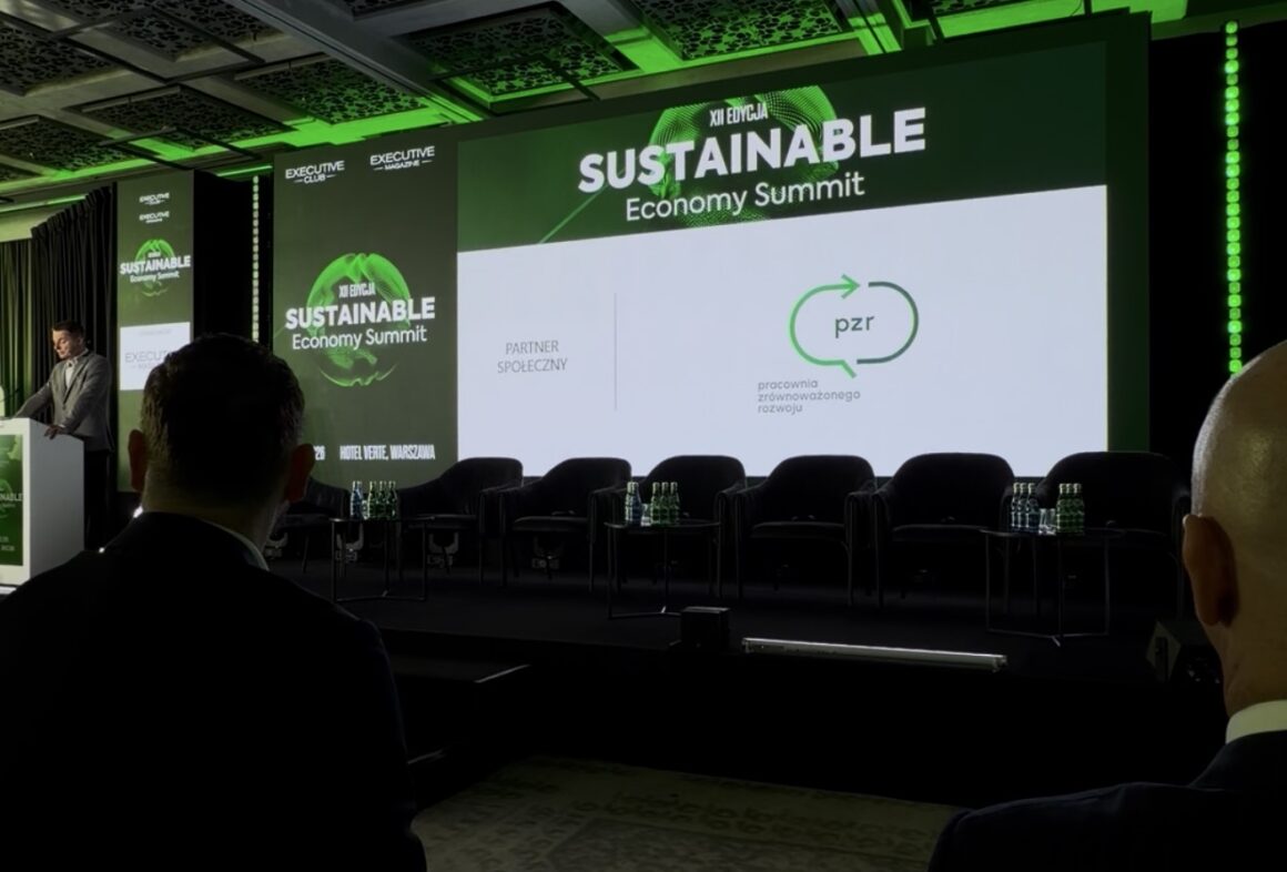 Zdjęcie przedstawia scenę konferencji Sustainable Economy Summit z dużym ekranem i zielonym oświetleniem. Na centralnym ekranie widoczny jest logotyp Pracowni Zrównoważonego Rozwoju jako partnera społecznego wydarzenia. Po lewej stronie osoba stoi przy mównicy, a przed sceną siedzą uczestnicy konferencji oglądający prezentację.