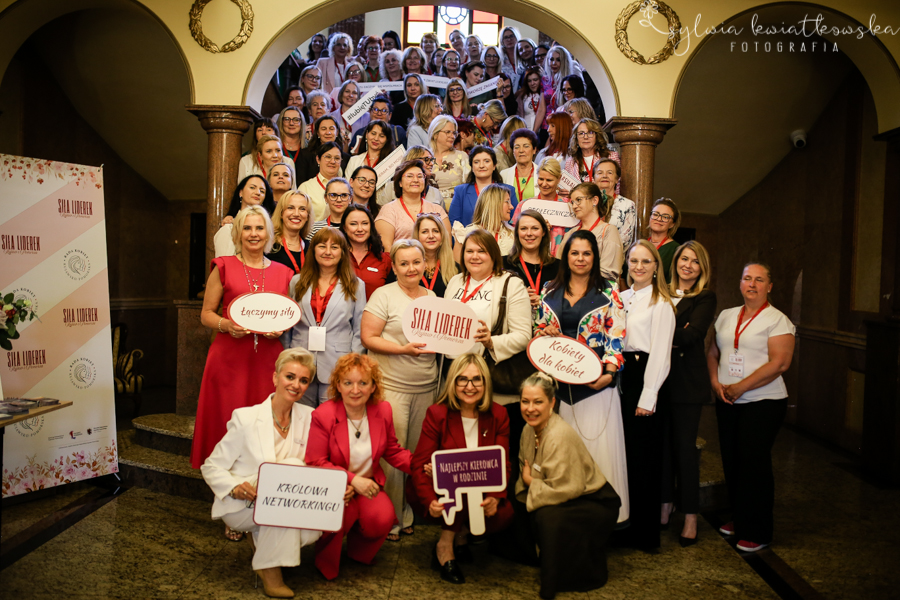 Duża grupa kobiet pozuje do wspólnego zdjęcia na reprezentacyjnych schodach wewnątrz zabytkowego budynku. Uczestniczki wydarzenia stoją na kilku poziomach schodów i uśmiechają się do aparatu, wiele z nich ma na szyjach identyfikatory. W pierwszym rzędzie kilka osób trzyma tabliczki z hasłami „Siła liderek”, „Łączymy siły”, „Kobiety dla kobiet”, „Królowa networkingu” oraz „Najlepszy kierownik w rodzinie”. Po lewej stronie widoczny jest roll-up wydarzenia „Siła Liderek”. Zdjęcie ma charakter pamiątkowy i pokazuje atmosferę integracji, współpracy oraz wzajemnego wsparcia kobiet uczestniczących w spotkaniu.
