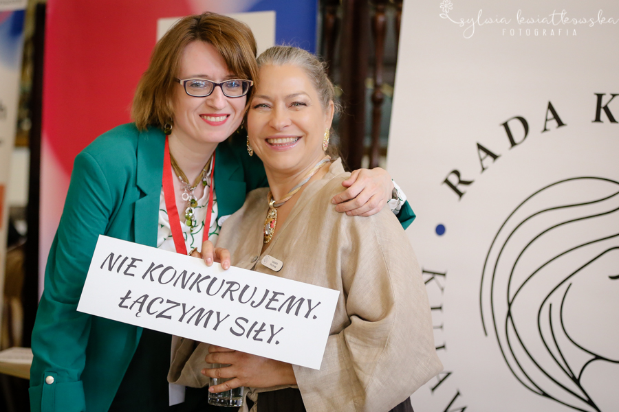 Dwie uśmiechnięte kobiety pozują do zdjęcia podczas wydarzenia „Siła Liderek Kujaw i Pomorza”. Stoją blisko siebie, obejmując się ramionami. Jedna z nich trzyma kartkę z napisem: „Nie konkurujemy. Łączymy siły.” W tle widoczne są elementy identyfikacji wizualnej wydarzenia oraz fragment logo Rady Kobiet Województwa Kujawsko-Pomorskiego. Zdjęcie podkreśla ideę współpracy, wzajemnego wsparcia i budowania relacji między aktywnymi kobietami.
