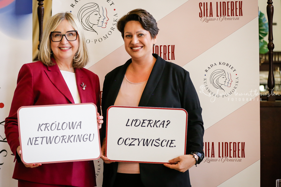 Dwie uśmiechnięte kobiety stoją przed ścianką promującą wydarzenie „Siła Liderek Kujaw i Pomorza”. Trzymają tabliczki z napisami „Królowa networkingu” oraz „Liderka? Oczywiście.” Obie patrzą w stronę aparatu i pozują do pamiątkowego zdjęcia. W tle widoczne są logotypy Rady Kobiet Województwa Kujawsko-Pomorskiego oraz grafiki związane z wydarzeniem. Zdjęcie podkreśla rolę kobiet liderek oraz znaczenie budowania sieci współpracy i wzajemnego wsparcia.