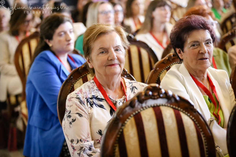 Dwie starsze uczestniczki wydarzenia siedzą w pierwszych rzędach sali i słuchają wystąpienia. Obie mają na szyjach czerwone identyfikatory konferencyjne i siedzą na ozdobnych, drewnianych krzesłach z tapicerowanymi oparciami. Jedna z kobiet patrzy bezpośrednio w stronę aparatu, druga zwraca uwagę na scenę. W tle widoczne są pozostałe uczestniczki spotkania. Zdjęcie pokazuje zaangażowanie kobiet w różnym wieku oraz międzypokoleniowy charakter wydarzenia „Siła Liderek Kujaw i Pomorza”.