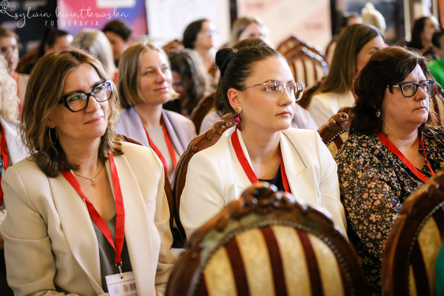 Trzy uczestniczki wydarzenia siedzą w pierwszym rzędzie sali i z uwagą słuchają wystąpienia. Wszystkie mają na szyjach czerwone identyfikatory konferencyjne. Kobieta po lewej stronie ma okulary i jasną marynarkę, środkowa uczestniczka ubrana jest w białą marynarkę i patrzy w stronę sceny z wyraźnym skupieniem, a kobieta po prawej ma ciemne włosy, okulary i wzorzystą bluzkę. W tle widoczne są kolejne osoby uczestniczące w spotkaniu. Zdjęcie pokazuje atmosferę zaangażowania i uważnego słuchania podczas wydarzenia „Siła Liderek Kujaw i Pomorza”.