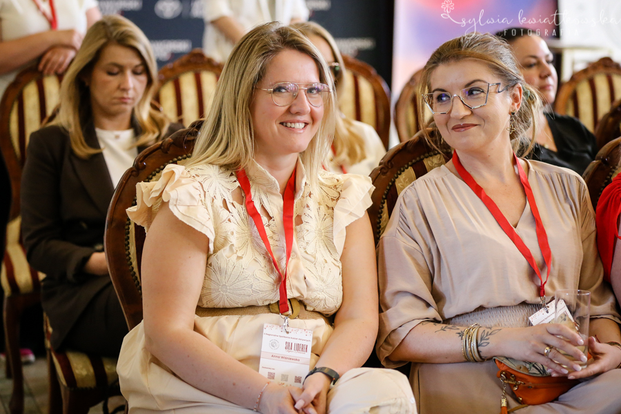 Dwie uczestniczki wydarzenia siedzą obok siebie w pierwszych rzędach sali konferencyjnej. Obie mają na szyjach czerwone identyfikatory uczestniczek. Kobieta po lewej stronie, ubrana w jasny, kremowy strój z ozdobnymi falbanami przy rękawach, uśmiecha się w stronę aparatu. Kobieta po prawej stronie, w jasnobeżowej bluzce i okularach, spogląda na swoją sąsiadkę z życzliwym uśmiechem, trzymając w dłoni kieliszek. W tle widać inne uczestniczki siedzące na ozdobnych krzesłach. Zdjęcie oddaje atmosferę życzliwości, relacji i wzajemnego wsparcia podczas wydarzenia „Siła Liderek Kujaw i Pomorza”.
