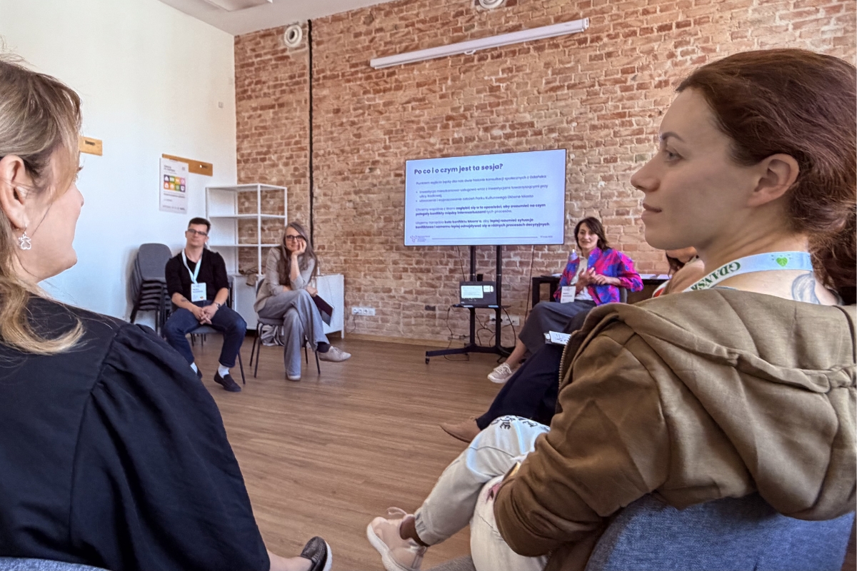 Uczestnicy i uczestniczki siedzą w kręgu podczas sesji warsztatowej na Regionalnym Forum Praktyków Partycypacji w Gdańsku. W tle widać ekran z prezentacją oraz ceglaną ścianę sali warsztatowej. Osoby rozmawiają o konfliktach i procesach podejmowania decyzji w działaniach partycypacyjnych.