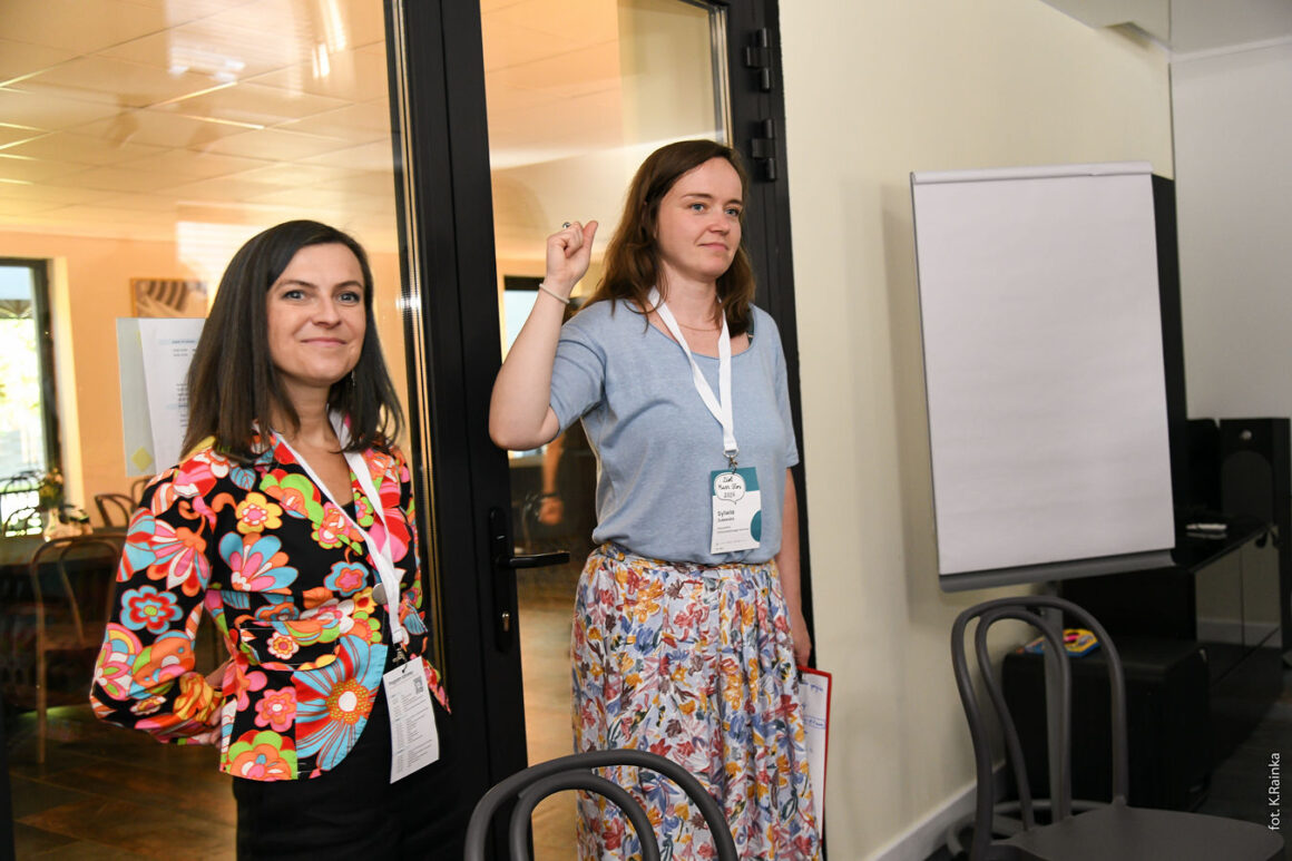 Dwie kobiety prowadzące warsztat stoją przy wejściu do sali konferencyjnej. Obie mają identyfikatory uczestniczek programu Masz Głos. Kobieta po lewej ubrana jest w kolorową bluzkę w kwiatowy wzór i uśmiecha się w stronę uczestników. Kobieta po prawej, w jasnoniebieskiej koszulce i kwiecistej spódnicy, unosi rękę, jakby udzielała wskazówki lub rozpoczynała ćwiczenie. Obok nich stoi pusty flipchart, a na pierwszym planie widać krzesła przygotowane do pracy warsztatowej. Zdjęcie przedstawia moment otwarcia lub prowadzenia szkolenia w kameralnej atmosferze.