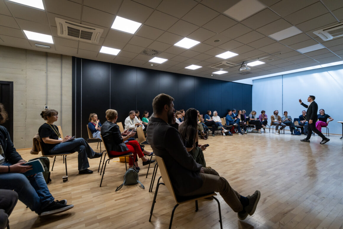 Szerokie ujęcie sali warsztatowej podczas Forum Praktyków Partycypacji. Uczestniczki i uczestnicy siedzą w dużym kręgu ustawionym z krzeseł, rozmawiają i słuchają prowadzącego. Po prawej stronie kadru prowadzący stoi na środku otwartej przestrzeni i zwraca się do grupy, angażując uczestników w ćwiczenie lub dyskusję. Jasna, nowoczesna sala z drewnianą podłogą i wysokim sufitem podkreśla otwartą formułę spotkania. Zdjęcie pokazuje atmosferę współpracy, aktywnego udziału i wspólnego uczenia się w gronie osób zajmujących się partycypacją obywatelską.