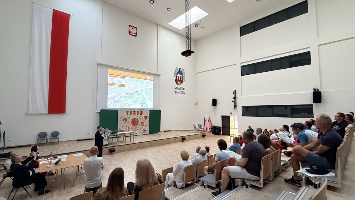 Spotkanie konsultacyjne dotyczące zmian w komunikacji miejskiej odbywające się w dużej sali widowiskowej. Na scenie prowadzący prezentuje slajd z mapą projektowanych linii komunikacyjnych, a uczestnicy siedzą na widowni i śledzą prezentację, część z nich trzyma w rękach materiały informacyjne. Na ścianach widoczne są flaga Polski, herb i logo Miasta Torunia. Zdjęcie przedstawia otwartą prezentację połączoną z konsultacjami społecznymi, podczas których mieszkańcy zapoznają się z propozycjami zmian i przygotowują do dyskusji.