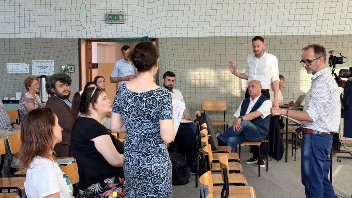 Spotkanie konsultacyjne odbywające się w sali gimnastycznej. Kilkanaście osób siedzi na krzesłach ustawionych w półokręgu i bierze udział w dyskusji dotyczącej komunikacji miejskiej. Na pierwszym planie jedna z uczestniczek stoi z materiałami w ręku i zabiera głos. Po prawej stronie prowadzący spotkanie odpowiada na pytania, a obok niego druga osoba notuje uwagi. W tle widoczne są kolejne osoby słuchające dyskusji oraz sprzęt do rejestracji wydarzenia. Zdjęcie przedstawia aktywny moment konsultacji społecznych, podczas których mieszkańcy wymieniają opinie i zgłaszają swoje propozycje.