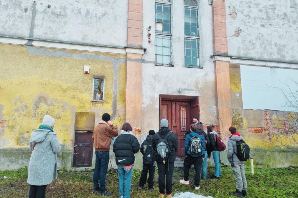 Grupa kilkunastu osób stoi przed wejściem do zabytkowego pałacu w Murowanej Goślinie podczas spaceru badawczego. Uczestnicy, ubrani w zimowe kurtki i z plecakami, słuchają osoby prowadzącej spotkanie stojącej przy głównych drzwiach budynku. Na zdjęciu widoczna jest zniszczona elewacja pałacu z odpadającym tynkiem, wysokimi oknami i śladami upływu czasu, co podkreśla potrzebę rewitalizacji obiektu. Zdjęcie dokumentuje terenowy etap procesu konsultacyjnego dotyczącego przyszłości pałacu.