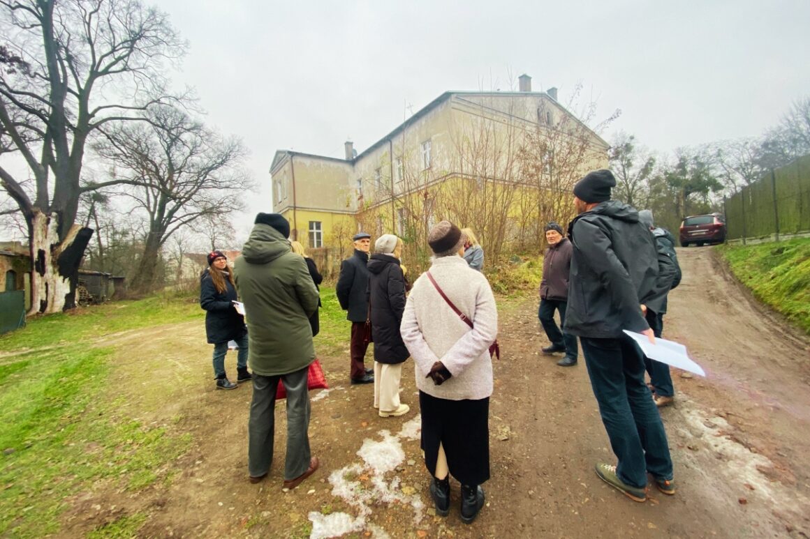 Grupa mieszkańców i uczestników procesu konsultacyjnego stoi przed pałacem w Murowanej Goślinie podczas spaceru badawczego. Osoby ubrane w zimowe kurtki rozmawiają i słuchają prowadzącego, trzymając w rękach notatki i materiały. W tle widoczny jest zabytkowy budynek pałacu otoczony drzewami i zielenią. Zdjęcie przedstawia terenowy etap diagnozy lokalnej, podczas którego uczestnicy wspólnie poznają otoczenie i dyskutują o przyszłości obiektu oraz jego najbliższego otoczenia.
