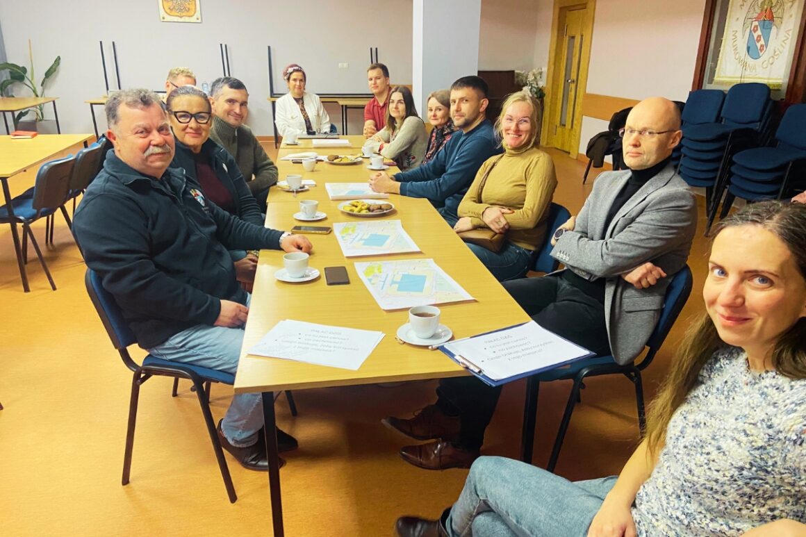 Grupa jedenastu osób siedzi przy dwóch połączonych stołach podczas warsztatu konsultacyjnego dotyczącego przyszłości pałacu w Murowanej Goślinie. Uczestnicy zwróceni są w stronę aparatu i uśmiechają się. Na stołach leżą mapy terenu, notatki, dokumenty, filiżanki z kawą i herbatą oraz talerze z poczęstunkiem. Spotkanie odbywa się w sali Urzędu Miasta i Gminy Murowana Goślina i ma roboczy charakter – uczestnicy wspólnie analizują materiały oraz rozmawiają o kierunkach zagospodarowania pałacu i jego otoczenia.
