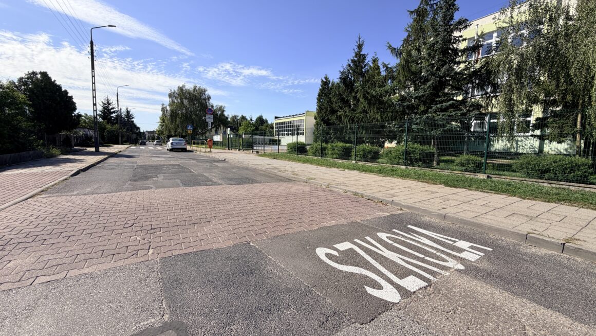 Zdjęcie ulicy Rzepakowej przy Szkole Podstawowej nr 9 w Toruniu. Po prawej stronie, za ogrodzeniem i drzewami, widoczny jest budynek szkoły. Na jezdni znajduje się próg zwalniający oraz duży biały napis „SZKOŁA”.