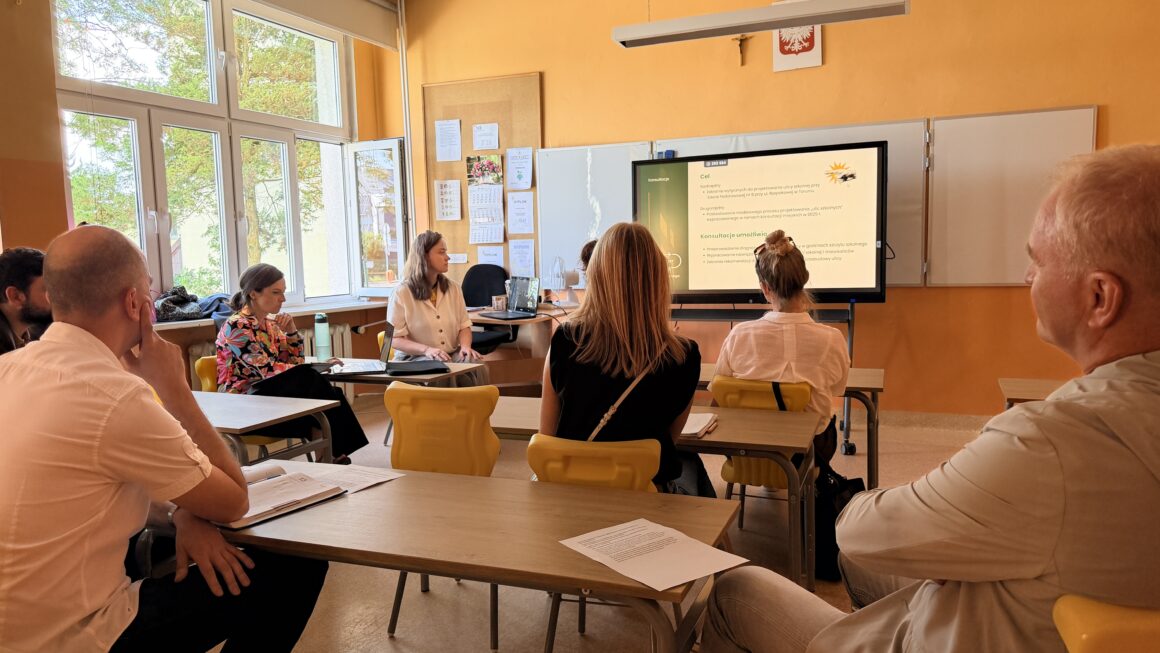 Zdjęcie ze spotkania przygotowawczego dotyczącego ulicy szkolnej przy Szkole Podstawowej nr 9 w Toruniu. Uczestnicy siedzą w szkolnej sali i słuchają prezentacji. Na ekranie wyświetlone są informacje o celu i planowanym przebiegu konsultacji.