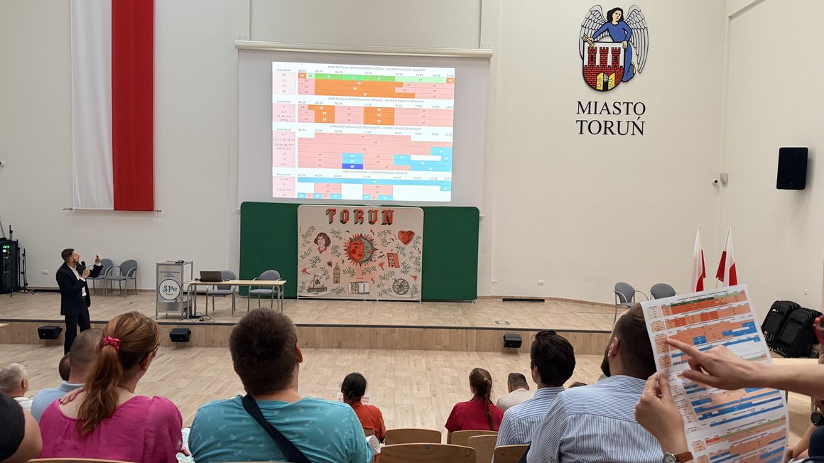 Zdjęcie ze spotkania dotyczącego konsultacji społecznych w sprawie zmian w komunikacji miejskiej w Toruniu. W dużej sali szkolnej mężczyzna w ciemnym garniturze przemawia do mikrofonu, wskazując na ekran z prezentacją przedstawiającą kolorowe schematy rozkładów jazdy autobusów. Na pierwszym planie siedzą uczestnicy spotkania, a jedna z osób trzyma wydrukowany projekt rozkładu jazdy. W tle widoczna jest scena z dekoracją przedstawiającą Toruń, godło miasta na ścianie oraz polskie flagi.
