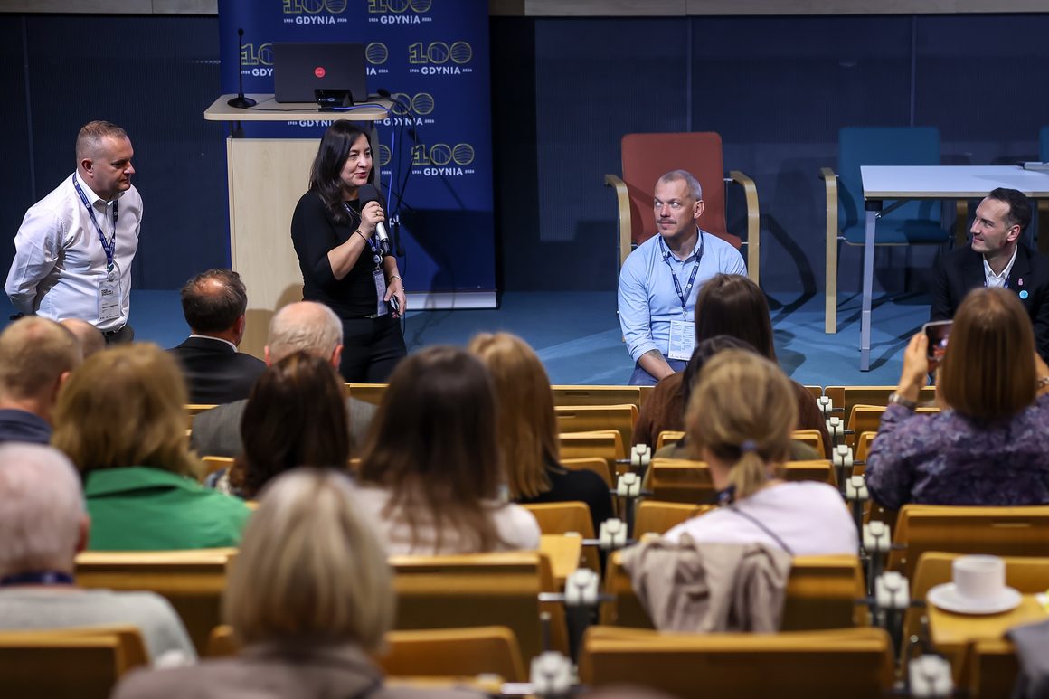 Zdjęcie z Forum Komitetów Rewitalizacji w Gdyni. W sali konferencyjnej kobieta ubrana na czarno stoi przed publicznością i przemawia do mikrofonu. Obok niej znajduje się mężczyzna w białej koszuli. Po prawej stronie dwóch mężczyzn, jeden w jasnoniebieskiej koszuli, drugi w ciemnej marynarce, przysłuchuje się wypowiedzi. Na pierwszym planie widoczni są uczestnicy siedzący w rzędach, zwróceni w stronę prelegentów. W tle znajduje się granatowa ścianka z logo 100-lecia Gdyni.