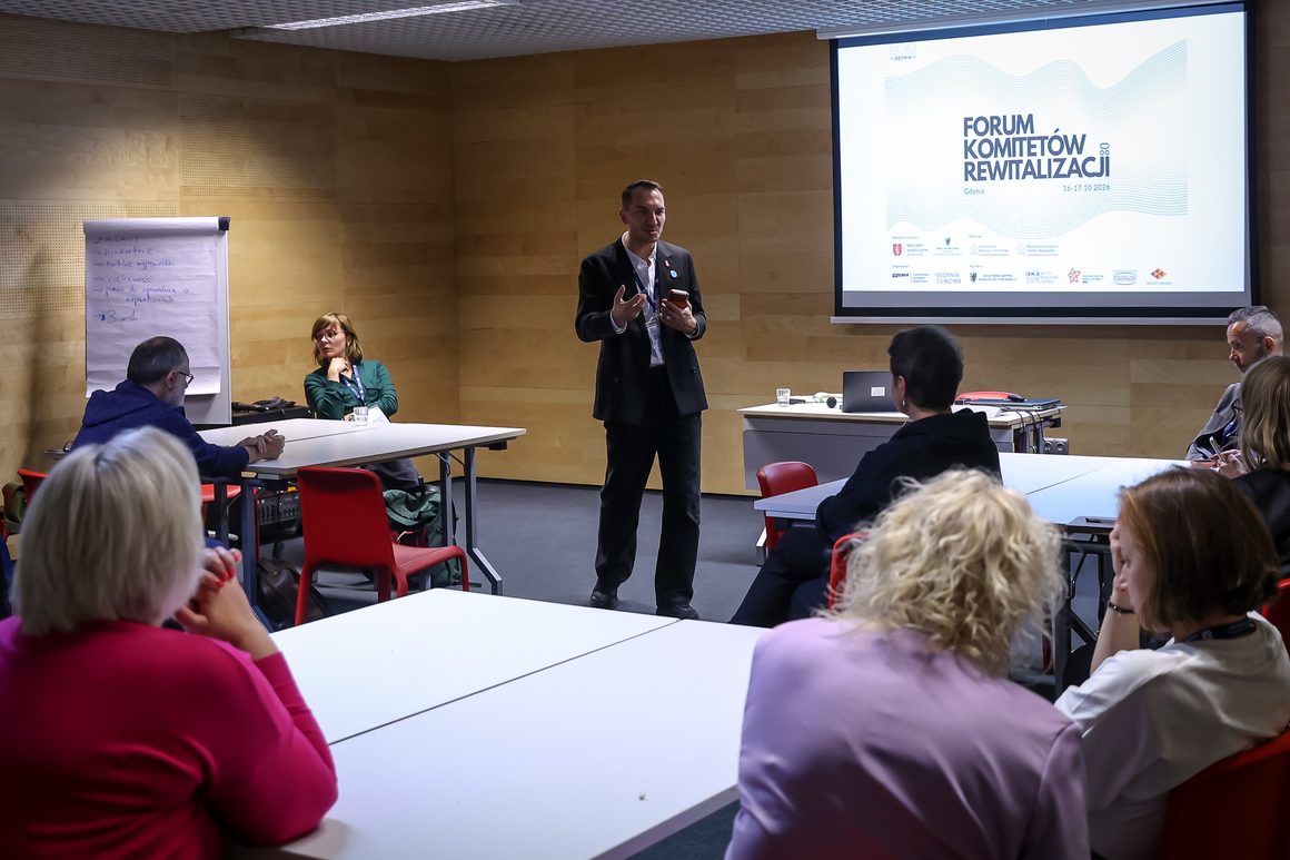 Zdjęcie z sesji warsztatowej podczas Forum Komitetów Rewitalizacji, które odbyło się 16–17 października 2026 roku w Gdyni. Na środku sali stoi mężczyzna w ciemnym garniturze, który prowadzi spotkanie, gestykulując i trzymając telefon w dłoni. Uczestniczki i uczestnicy siedzą przy stołach ustawionych w podkowę i słuchają prowadzącego. Po lewej stronie widoczna jest tablica z zapisanymi zasadami pracy, a na ekranie projekcyjnym znajduje się logo Forum Komitetów Rewitalizacji.