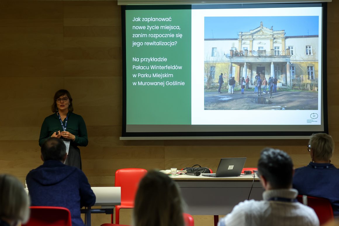 Zdjęcie z Forum Komitetów Rewitalizacji w Gdyni, 16–17 października 2026 roku. Po lewej stronie stoi kobieta w zielonej koszuli i okularach, prowadząca prezentację przed uczestnikami spotkania. Na dużym ekranie wyświetlony jest slajd Pracowni Zrównoważonego Rozwoju zatytułowany „Jak zaplanować nowe życie miejsca, zanim rozpocznie się jego rewitalizacja?”. Prezentacja dotyczy Pałacu Winterfeldów w Parku Miejskim w Murowanej Goślinie. Obok tekstu znajduje się zdjęcie zabytkowego pałacu z grupą osób stojących przed wejściem. Na pierwszym planie widoczni są uczestnicy siedzący przy stołach i słuchający wystąpienia.