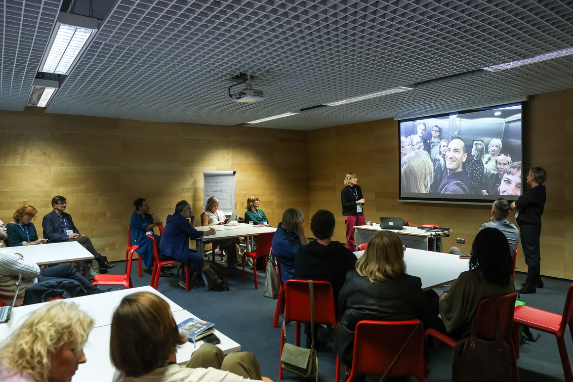 Zdjęcie z warsztatu podczas Forum Komitetów Rewitalizacji w Gdyni. W sali konferencyjnej kilkanaście osób siedzi przy stołach ustawionych w podkowę. Po prawej stronie dwie kobiety prowadzą prezentację. Na dużym ekranie wyświetlone jest grupowe zdjęcie uczestników wykonane w windzie. Jedna z prowadzących stoi przy ekranie, druga pośrodku sali. Uczestnicy obserwują prezentację i słuchają wystąpienia.